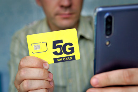 man holds a yellow SIM card with the inscription 5G and a mobile phone in his hand. Replacing the SIM card and switching to high-speed Internet.の写真素材