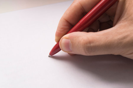 A mans hand writes with a red pen on a white paper surface. Top view of female hand with pencil on blank paper sheet. On cardboard backgroundの写真素材