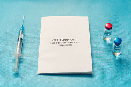 A document for recording vaccinations made in Russia, a syringe and a bottle of vaccine on a blue background. Translated from Russian: Certificate of preventive vaccinations.の写真素材