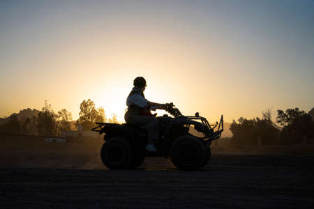 Silhouette of a tourist woman rides a quad bike against the background of a bright sunset. Evening extreme entertainment in the desert of Egypt.の写真素材