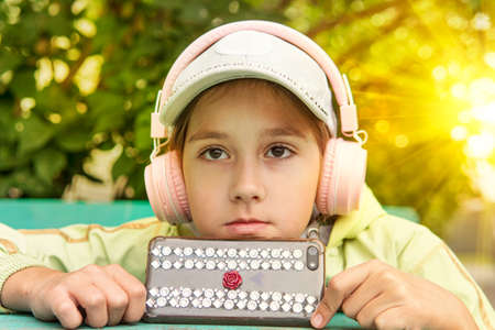 Portrait of beauty kid girl with long brunette hair wearing white cap with headphones listening to music outside. The child listens to audiobooks on the phone. Language training.の写真素材