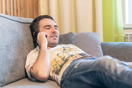 Caucasian man lie relax on sofa at home use modern smartphone gadget, relaxed millennial male rest...