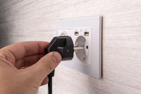 Hand plugging in an electric cord into a white plasticmodern socket or european wall outlet on gray wall. Closeup of a man hand inserting an electrical plug into a wall socket.の写真素材