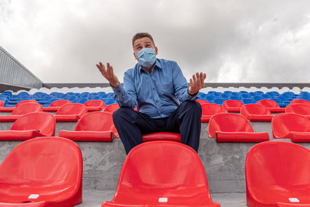lonely man with a protective mask on his face is sitting and watching a sports competition alone on a large empty grandstand. Competition and other mass events during the lockdown.の写真素材