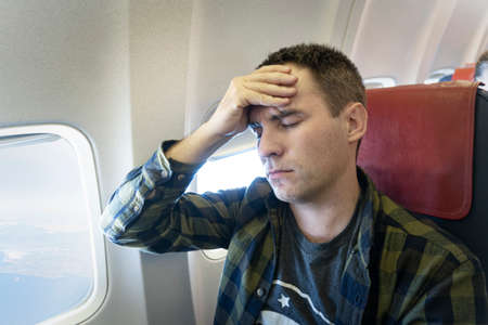 Frustrated man touching his head with hands and keeping eyes closed while sitting at his seat airplane. concept of aerophobia and high pressure while flying in airplane. health problems. Hard flight.の写真素材