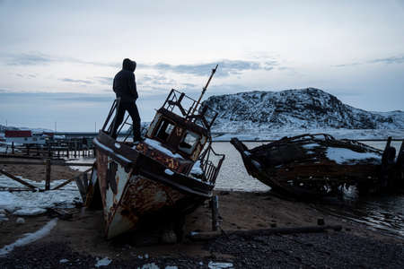a male tourist stands on a destroyed ship on the shore of the Barents Sea. The village of Teriberka,...