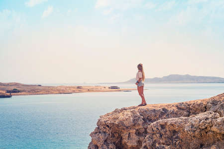 Rear view of young woman standing on mountainous road and admiring beautiful landscape of sea and mountains. tourist girl walking on trail in summer day. Travel destinations, staycation concept.の写真素材