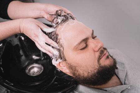 A young bearded man of Caucasian appearance at a reception at a barber shop. The process of cutting hair. Hairstylist washing clients hair in barber shopの写真素材