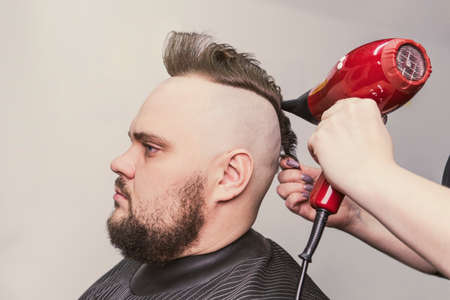 Female barber makes a mohawk hairstyle at adult man with beard. hair styling with a hair dryer.