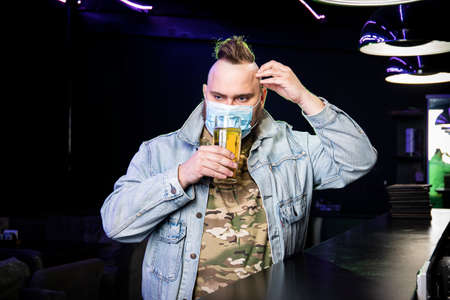 punk man with a mohawk wearing a protective mask on his face drinks beer from a glass mug in the dark. Quarantine in the bar. Prohibition of alcohol consumption. The concept of a new normality.の写真素材