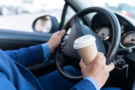 Man driver hand holding take away go coffee cup while driving a car in the morning, close up, people lifestyle, summer holiday journey concept. dangerous driving with a cup of coffeeの写真素材