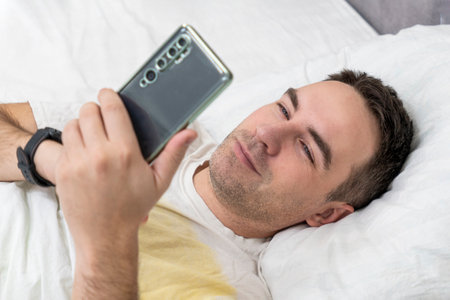 Joyful man woke up in the morning in his bedroom, uses smart phone, top view. A man smiles in bed...