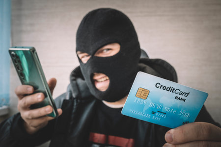 Close up of a man in robbery mask and hood holding the credit card and looking at the smartphone...