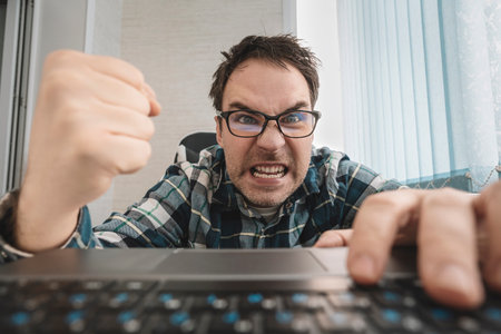 angry man swearing and cursing against information technology and his computer worries and hassles - concept of hating computers. businessman in a shirt in the office punches the keyboard.の写真素材
