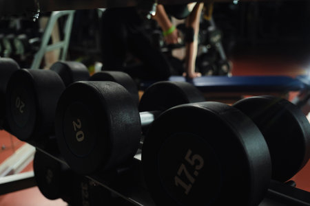 heavy black metal dumbbells for strength training on the rack. a woman lifts a dumbbell in the background. lifting dumbbellsの写真素材
