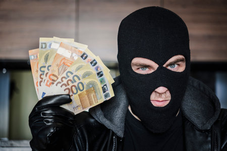 Male thief with balaclava on his head holding a handful of euro bills in front of his eyes. the...