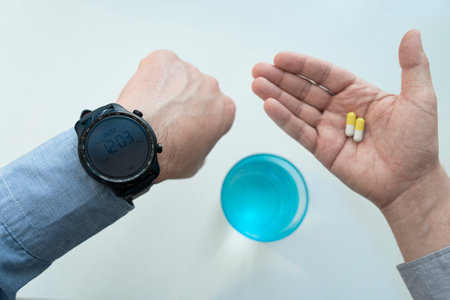 a businessman man in the office is looking at his watch and holding yellow pills in his hand on a white background. The concept of timely medication intake. treatment time.の写真素材
