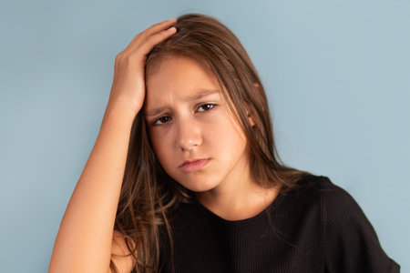 Little tired exhausted kid teen girl of 10-12 years old in black T-shirt put hand on forehead. look camera on pastel plain light blue background..の写真素材