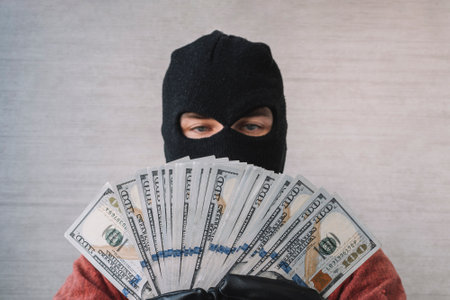 Male thief with balaclava on his head holding a handful of dollar bills in front of his eyes. the concept of crime and money theftの写真素材