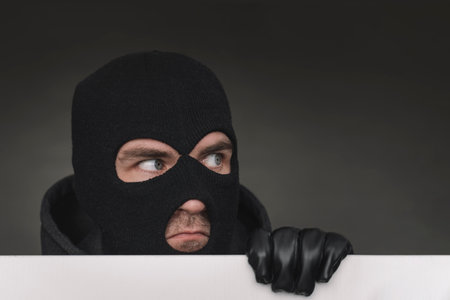 An evil thief in a mask looks out from behind a white board. Space for text and design. copy space. the surprised bandit looks awayの写真素材