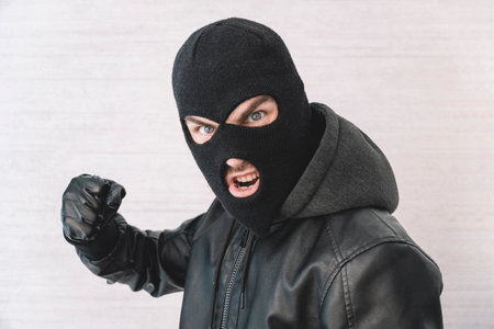 A thief in a black jacket. A man in a black balaclava with an evil expression on his face. An aggressive bandit on a white background. The concept of crime or theft.の写真素材
