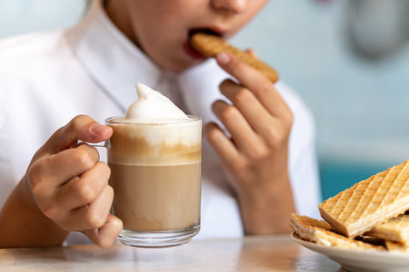 a young girl is eating a waffle and drinking cappuccino coffee with white foam. . Morning coffee and sweets in hands.の写真素材