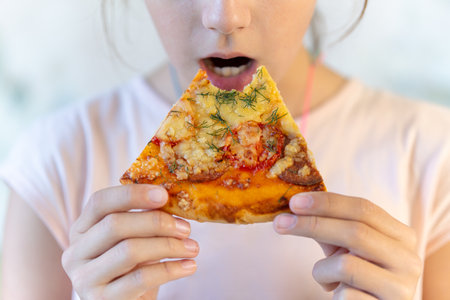 Teenage girl eats pizza. girl holds a pizza in his hands and eats at a sidewalk cafe. Street food and fast food concept. Unhealthy diet as a cause of gastritis diseases.の写真素材