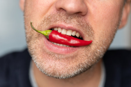 Expressive and funny man holding a whole red chili pepper in his mouth. Man eating a spicy pepper on a blue background. Extra hot cayenne pepper.の写真素材