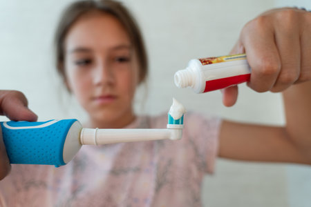 A young girl squeezes toothpaste on to electric toothbrush in a close-up in the bathroom. Brushing...