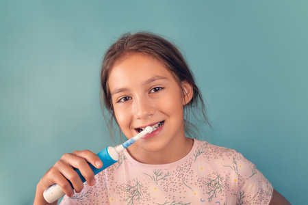 Beauty portrait of happy cheerful positive beautiful girl, young teen is brushing her healthy white teeth with electric toothbrush with toothpaste, looking at camera and smiling on blue background.の写真素材
