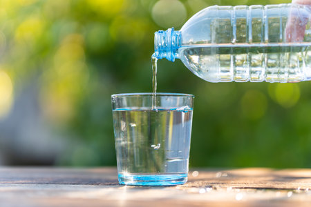 Hand holding drinking water bottle pouring into glass on wooden table on blurred green nature...
