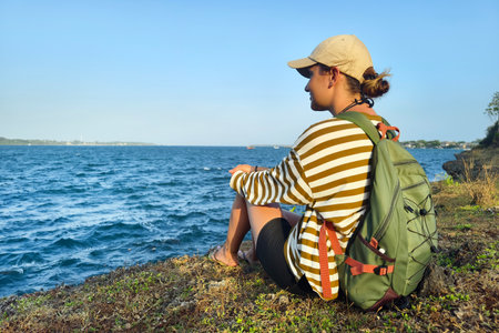 rear view Young woman wearing cap with backpack sitting on rocks and looking at view of ocean....