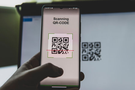 young businessman sitting at his workplace in front of computer monitor and scanning QR code with mobile smart phone.の写真素材