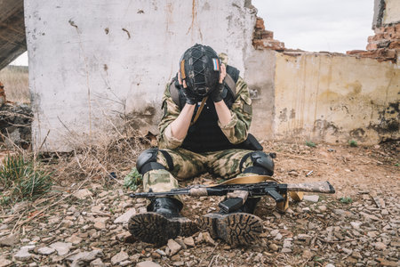 Post-traumatic stress disorder concept. A soldier in uniform with a machine gun sits on the ruins of a destroyed building and covers his face with his hand.の写真素材