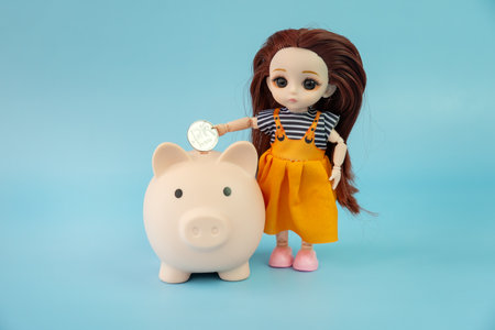 A small child doll with a coin in her hands on a blue background. The doll puts a coin in the piggy bank.の写真素材