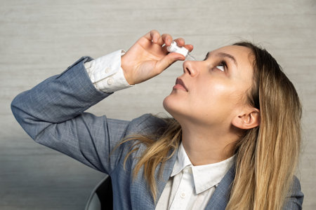 Female entrepreneur freelancer working on laptop and holding medicinal eye drops to relief eye strain and dryness, due to overwork on laptop. Digital eye strain. Ophthalmology. Healthcare and medicineの写真素材