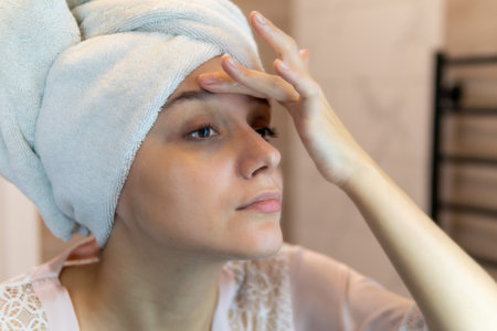 Young caucasian blonde woman looking in the bathroom mirror at her eyes with dark circles under the eyes. Bruises are caused by insomnia, fatigue, nervousness, lack of sleep and stressの写真素材