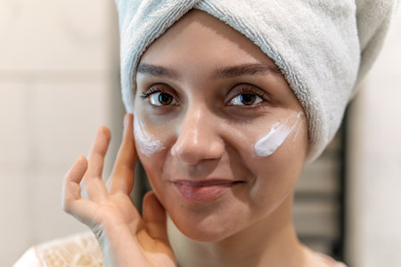 girl in the bathroom with a towel on her head and. spa treatments in front of a mirror, body care for a beautiful woman, girl smiling takes care. woman with a towel on her head in front of bathroom mirrorの写真素材