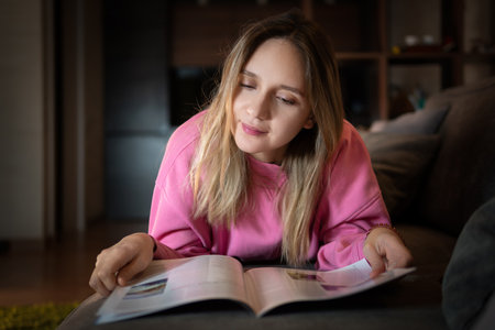 Happy woman reading a magazine lying on a sofa in the living room at home. A blonde girl is relaxing at home in the living room.の写真素材