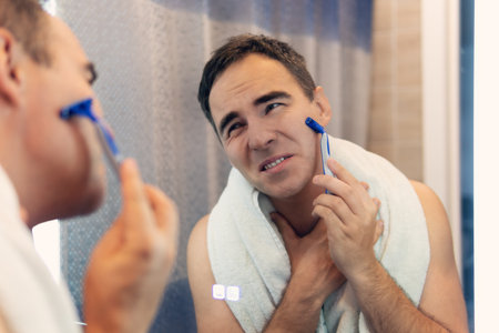 A man shaves his face without cream or foam. Man shaves in front of a mirror in the morning. Shaving of the face with skin irritation.の写真素材