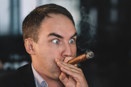 a man smokes a cigar, inhaling smoke. The harm of smoking is a concept. A guy with wide-open eyes inhales smoke.の写真素材