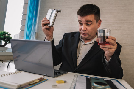 A businessman holds a flask and a glass while sitting at a table next to a laptop in an office.の写真素材