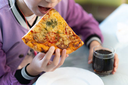 Teenage girl eats pizza. girl holds pizza in his hands and eats at sidewalk cafe. Street food and fast food concept. Unhealthy diet as a cause of gastritis. harmful food and harmful drinks.の写真素材