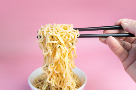 male hand stirs instant noodles with chopsticks in a bowl on a pink background.の写真素材