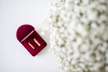 Wedding rings in a red velvet box and bridal bouquet over a white table - selective focus, copy...