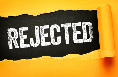 REJECTED text in bold, distressed white font revealed beneath torn yellow paper on a dark black background. Concept of denial, failure, disapproval, rejection, and negative decisionの写真素材