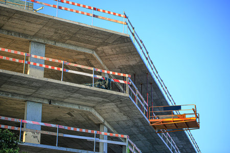 Modern concrete building under construction with orange suspended lift platform and red safety...