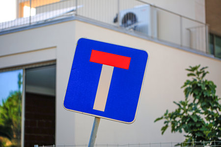 Close-up of a dead-end road sign (no through road) with a modern residential building and foliage in the background. The tilted sign stands out against the urban setting and neutral facadeの写真素材