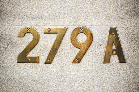 House number 279A in vintage brass mounted with visible screws on a lightly textured stone wall. The weathered patina and subtle rust give the numbers a rustic, aged look against the neutral backgroundの写真素材
