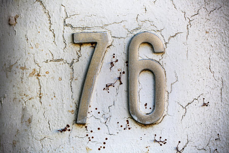 Metal house number 76 mounted on a white wall with peeling paint and rust stains. The textured...